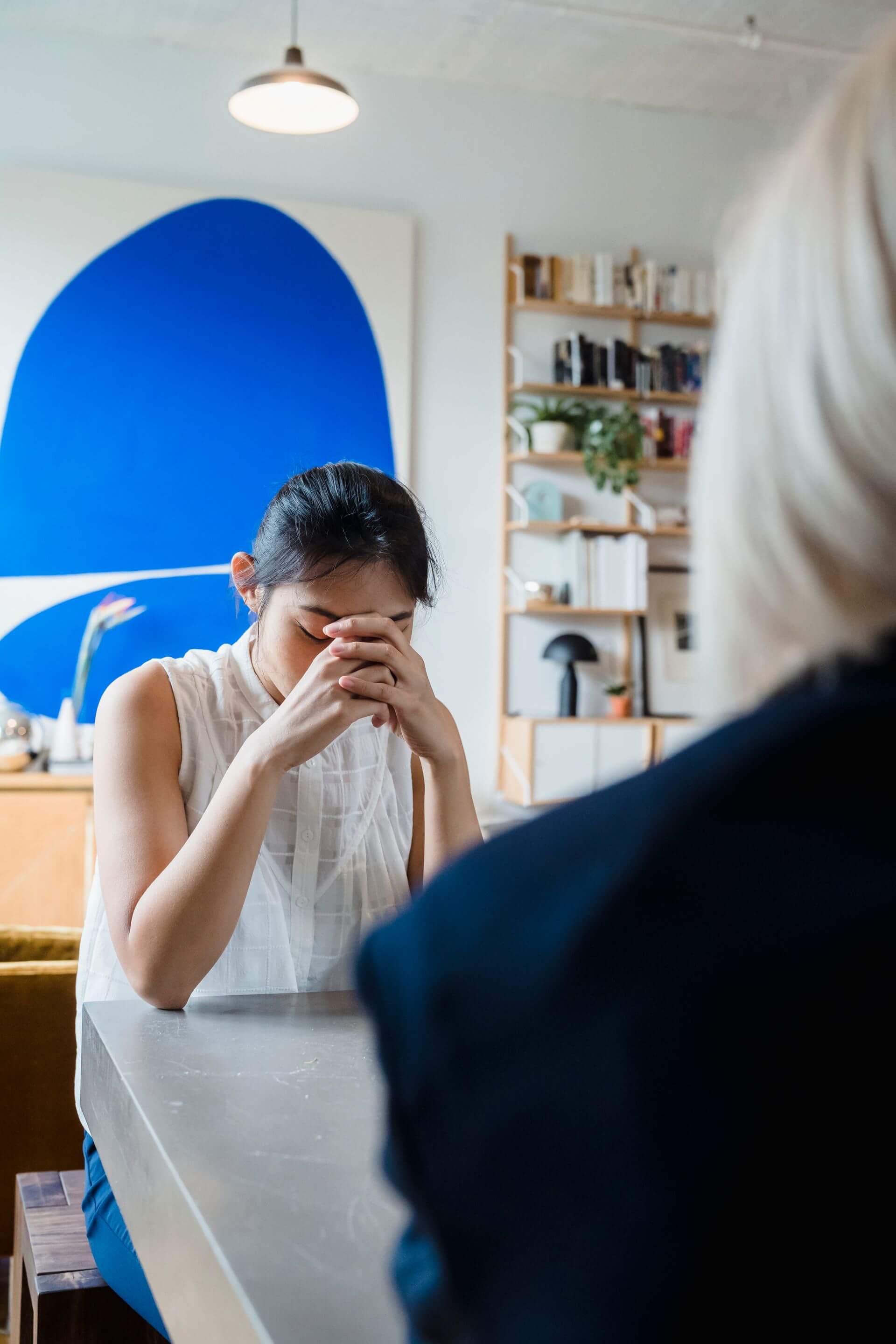 A woman sitting with her head bowed and hands clasped in a quiet moment of reflection, with her therapist nearby