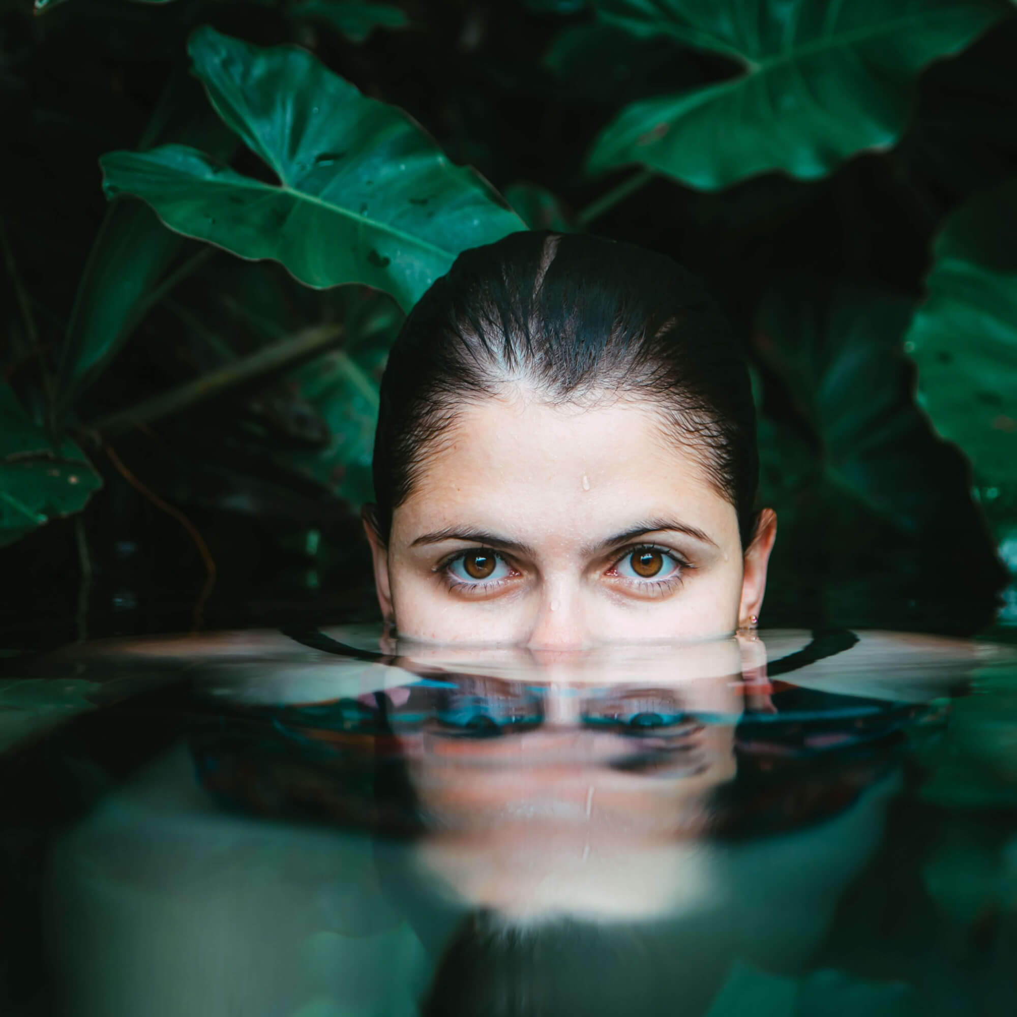 A woman's face partially submerged in still water, her reflection visible on the surface